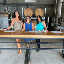 Load image into Gallery viewer, Three ladies dressed up in their best summer apparel pose for a picture, standing in the cellar and tasting room of Terravista vineyards on the Naramata Bench in Penticton, BC.  | Farm to Glass Wine Tours
