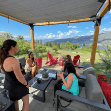 Load image into Gallery viewer, Three guests of Farm to Glass Wine Tours sit under a shaded gazebo tasting wine while owner and host, Michelle Sewchuck of Black Market Wine Co in Kaleden, BC explains how the wine was made.  The back drop of Skaha moutains in the background have shadows casted by the clouds and Skaha Lake with rolling vineyards in the background.  | Farm to Glass Wine Tours