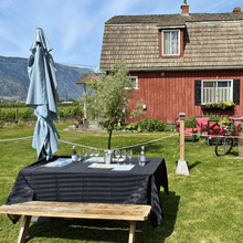 Load image into Gallery viewer, A picnic table set up for a premium wine tasting is ready for guests at Clos du Soleil Estate Winery in Keremeos, BC in the Similkameen Valley.  A red bard with black window shutters stands tall in the background with vineyards in view in the far distance and a steep mountain backdrop that is signature to this valley.  | Farm to Glass Wine Tours