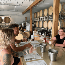 Load image into Gallery viewer, A couple leans into the tasting room bar at Anthony Buchanan Wines in Kaleden, BC while Tasting Room Manager Nikki Bailey explains the minimal intervention ways they make their wines.  In the background, stacks of barrels with the current vintage of wines await their turn to be bottled.  | Farm to Glass Wine Tours