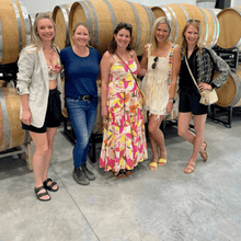 Load image into Gallery viewer, Four best girlfriends strike a pose, smiling in front of wine barrels at Solvero Wines. The cellar's warm ambiance complements their joyous camaraderie. Winemaker Alison Moyes stands among them, sharing the passion for her craft. A delightful moment of friendship, wine, and shared experiences at Solvero Wines | Farm to Glass Wine Tours