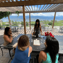 Load image into Gallery viewer, Seated at a long wooden table underneath a shaded gazebo, 3 guests look towards the host of Lunessence winery in Summerland, BC as she explains the wines in their wine tasting.  The gazebo sits on an edge that looks out onto the rolling vineyards and trees with Okanagan Lake and moutains in the background.  | Farm to Glass Wine Tours