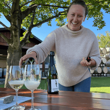 Load image into Gallery viewer, Tasting Room Manager Katrice Sutherland smiles and looks towards the camera as she pours two glasses of Lightning Rock Blanc de Noir sparkling wine on a picnic table.  Wine barrels linger in the background against a contrasting blue sky and large oak tree.  | Farm to Glass Wine Tours
