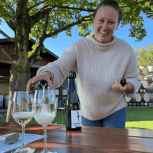 Load image into Gallery viewer, Tasting Room Manager Katrice Sutherland smiles and looks towards the camera as she pours two glasses of Lightning Rock Blanc de Noir sparkling wine on a picnic table.  Wine barrels linger in the background against a contrasting blue sky and large oak tree.  | Farm to Glass Wine Tours