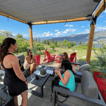 Load image into Gallery viewer, Three guests of Farm to Glass Wine Tours sit under a shaded gazebo tasting wine while owner and host, Michelle Sewchuck of Black Market Wine Co in Kaleden, BC explains how the wine was made.  The back drop of Skaha moutains in the background have shadows casted by the clouds and Skaha Lake with rolling vineyards in the background.  | Farm to Glass Wine Tours
