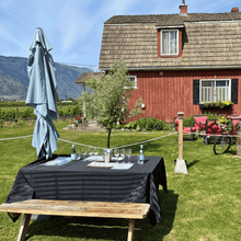 Load image into Gallery viewer, A picnic table set up for a premium wine tasting is ready for guests at Clos du Soleil Estate Winery in Keremeos, BC in the Similkameen Valley.  A red bard with black window shutters stands tall in the background with vineyards in view in the far distance and a steep mountain backdrop that is signature to this valley.  | Farm to Glass Wine Tours
