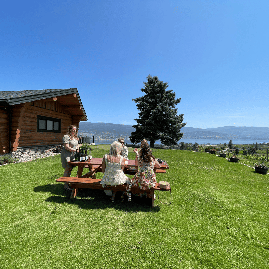 Four guests sit at a picnic table on a massive green lawn where Katrice Sutherland of Lightning Rock Winery in Summerland, BC guides them through a premium tasting outdoors under a bright blue sky.  Off in the distance is Okanagan Lake.  | Farm to Glass Wine Tours