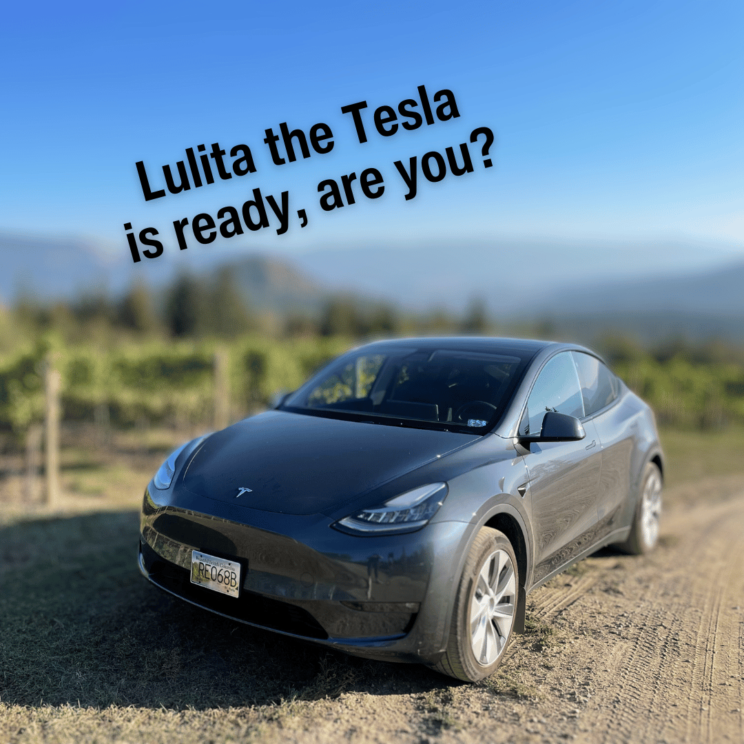 A sleek, grey Tesla electric car parked amidst scenic vineyards in the Okanagan Valley and at the top of the photo there is a caption that says, "Lulita the Tesla is ready, are you?".  This electric vehicle represents Farm to Glass Wine Tours, offering a luxurious and environmentally conscious way to explore the wine country of the Okanagan Valley