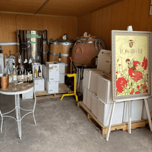 Load image into Gallery viewer, An easel with a poster of the logo of Fox &amp; Archer wines leans against boxes filled with cases of wine in a small garage that has been converted into a winery where a winery tank, a barrel and a clay barrel sit in the back.  There is a table with a lineup of wines from Fox &amp; Archer with wine glasses and a spit bucket ready for the next guests to arrive for a tasting.  | Farm to Glass Wine Tours.
