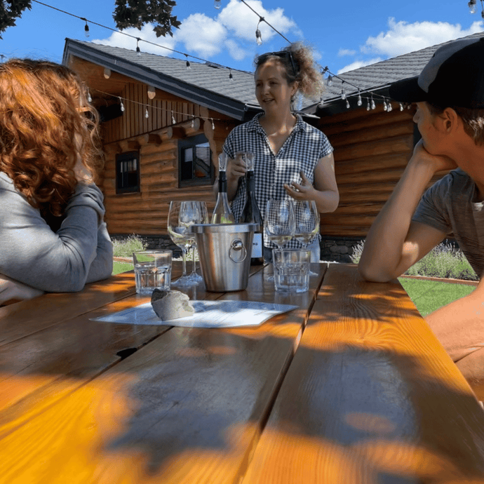 Winemaker Jordan Kubek, stands at the edge of a picnic table that has wine glasses and 2 bottles of Pinot Noir, water glasses while a couple seated look at Jordan, encaptured by the way Jordan explains how their Pinot Noir is made.  | Farm to Glass Wine Tours
